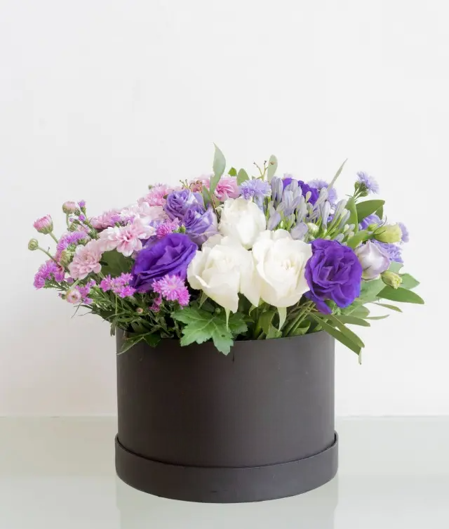 A flower arrangement with purple , white roses with chrysanthemum in a black round box