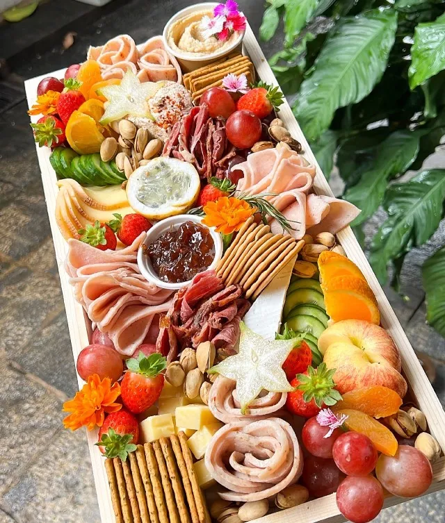 This platter serves up to 6 people with a variety of flavors. It includes five cheeses: Smoked Scamorza, Cheddar, Herb & Garlic Neufchatel, Chili Pepper Neufchatel, and Pistachio-Cranberry Cheese Ball. Paired with cold cuts like Beef Mortadella, Chicken Ham, Smoked Beef, and Chicken Mortadella, it also features fresh fruit, nuts, olives, crackers, hummus dip, chutney, and a dried orange and rosemary garnish for a perfect balance of savory, sweet, and tangy.