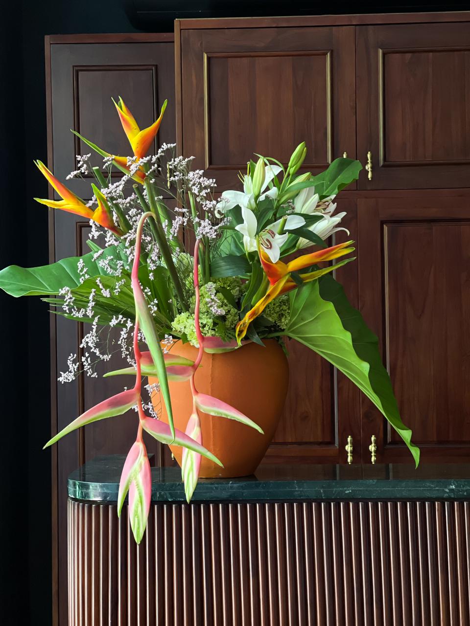 Surprise your loved one with an exquisite flower arrangement featuring vibrant heliconias, alluring Sexy Pink heliconias, delicate lilies, and lush hydrangeas, all elegantly presented in a stunning orange pot. A gift of beauty and grace that speaks volumes