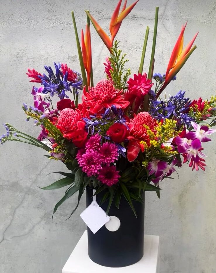 A striking tropical arrangement, beautifully presented in a sleek black basket. This vibrant display features bold heliconias, exotic orchids, and soft chrysanthemums, creating a perfect blend of color and elegance.