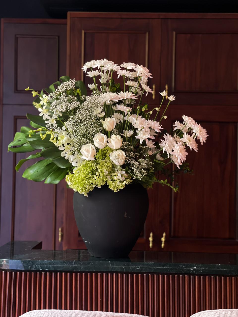 Gift a sophisticated arrangement of white roses, lush hydrangeas, delicate chrysanthemums, and elegant orchids, accented with fresh foliage in a sleek black vase. A timeless expression of beauty and grace for any occasion.