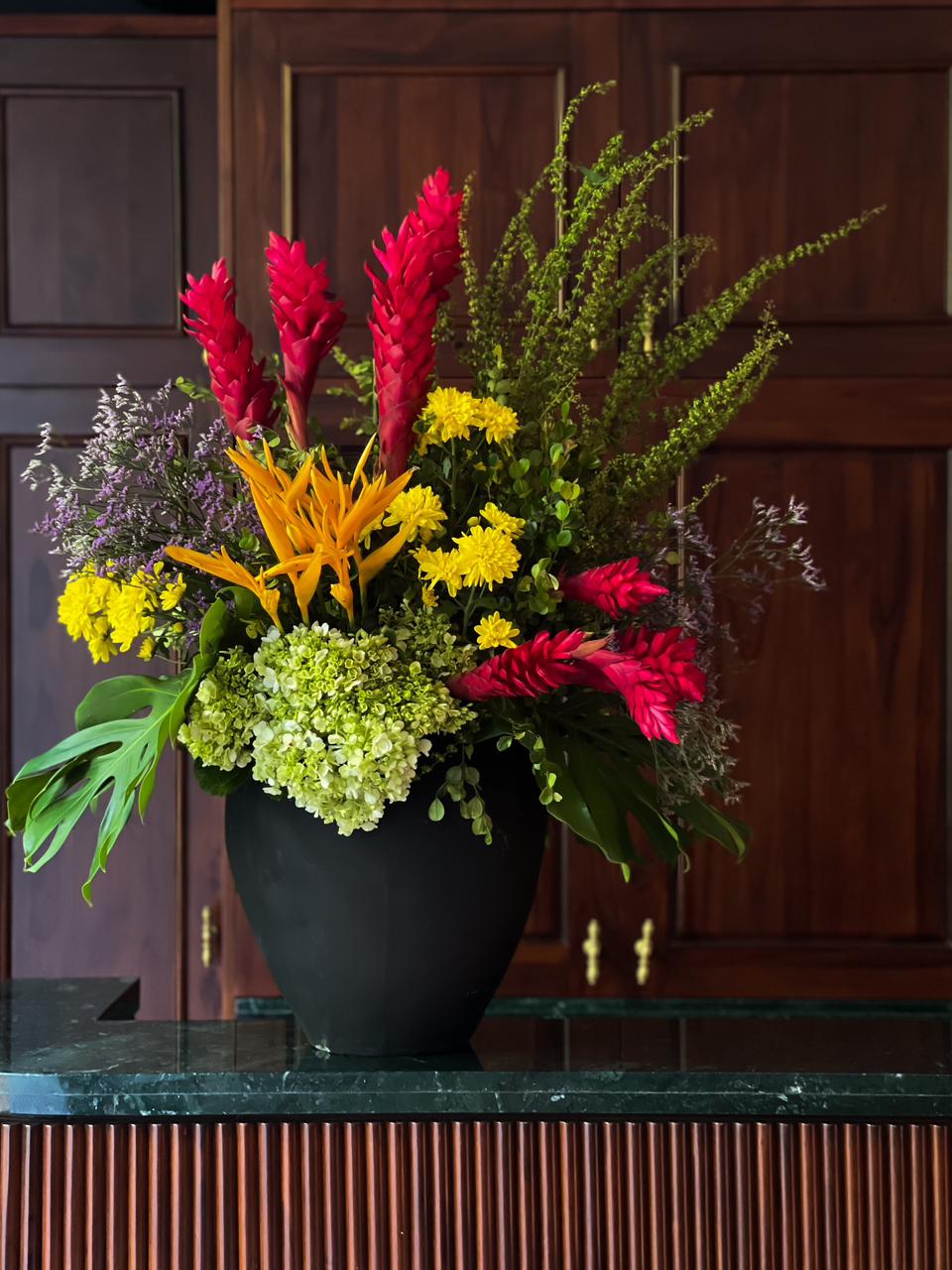 Gift a stunning arrangement of vibrant tropical flowers, lush hydrangeas, and delicate chrysanthemums, accented with exotic foliage and elegantly presented in a sleek black vase. A perfect blend of beauty and sophistication for any occasion
