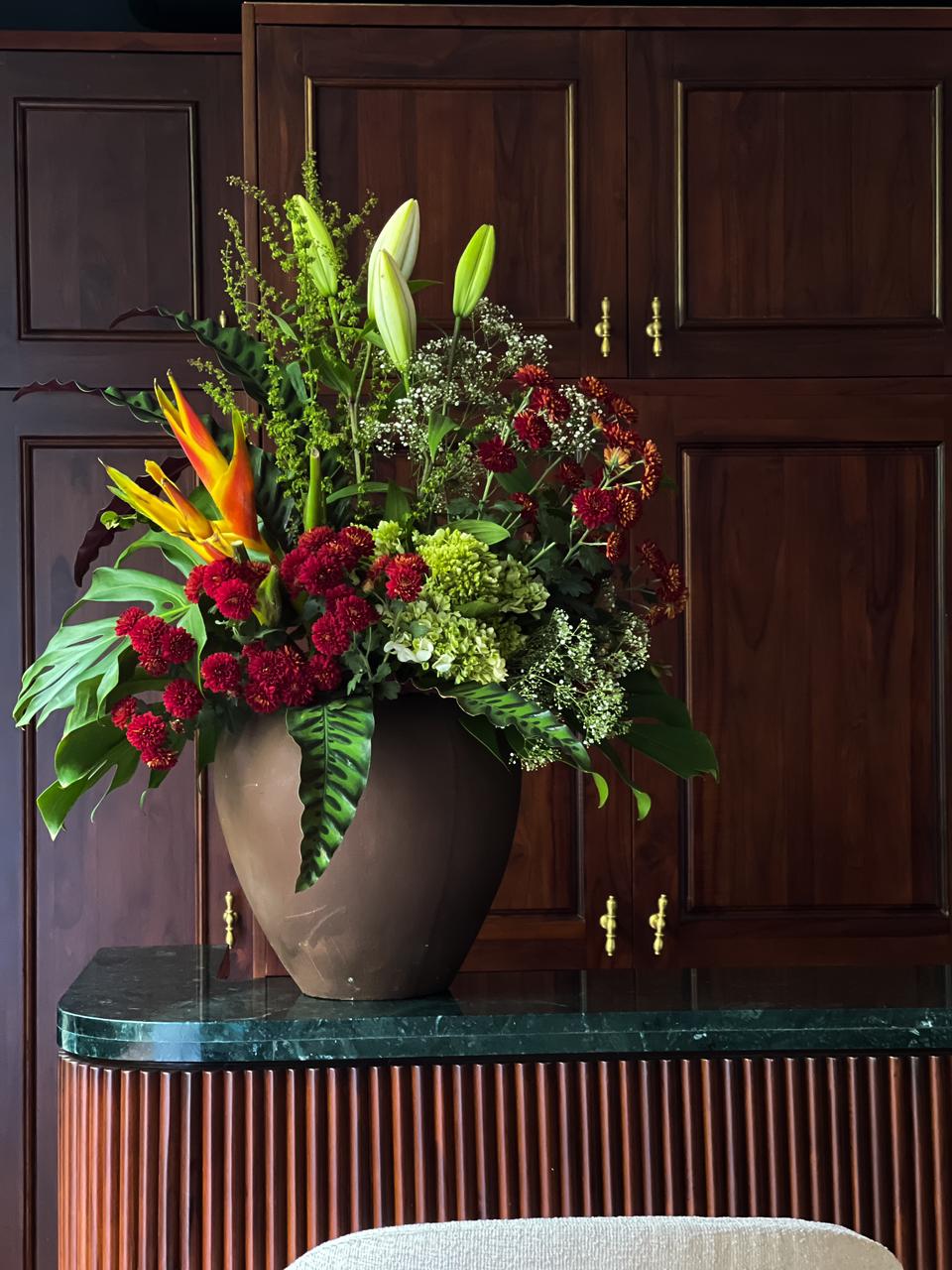 Gift a striking arrangement of maroon chrysanthemums, lush hydrangeas, exotic tropical flowers, and elegant lilies, accented with lush foliage in a sophisticated brown vase. A perfect statement of beauty and elegance for any occasion.